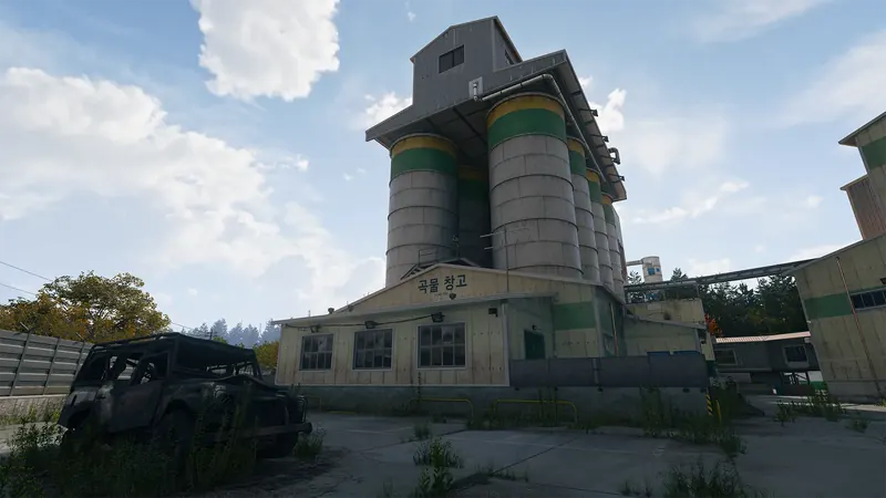 Tall concrete grain silos rise above a rice mill warehouse, with a burned-out vehicle parked in the foreground.